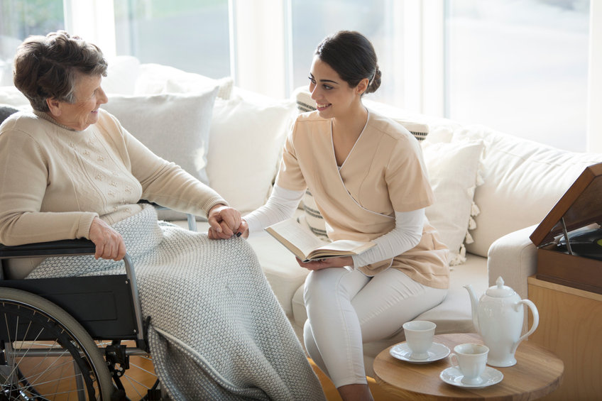 A disabled old woman in a wheelchair holding the hand of a tender professional medical assistant during tea time in a living room of luxury retirement home